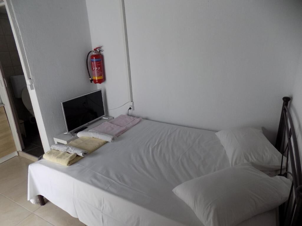 A neatly made bed with white linens and pillows, a bedside table with a flat-screen TV and a pink towel, and a fire extinguisher mounted on the wall.