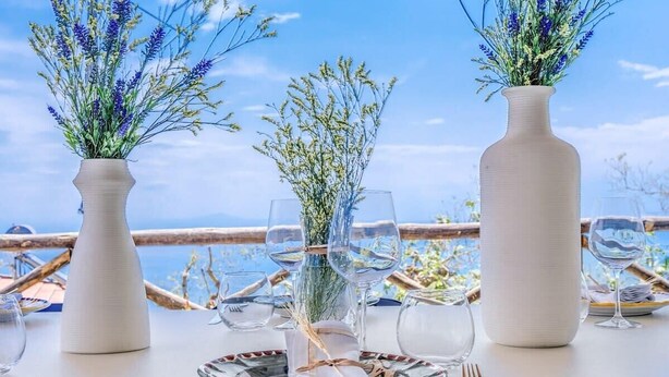 Une table dressée pour le dîner, ornée de vases blancs contenant des fleurs, de verres à vin transparents et de couverts, avec une vue panoramique en toile de fond.