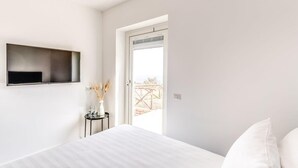 Une chambre avec un lit, un téléviseur fixé au mur, une petite table avec un vase et une porte donnant sur un espace extérieur.