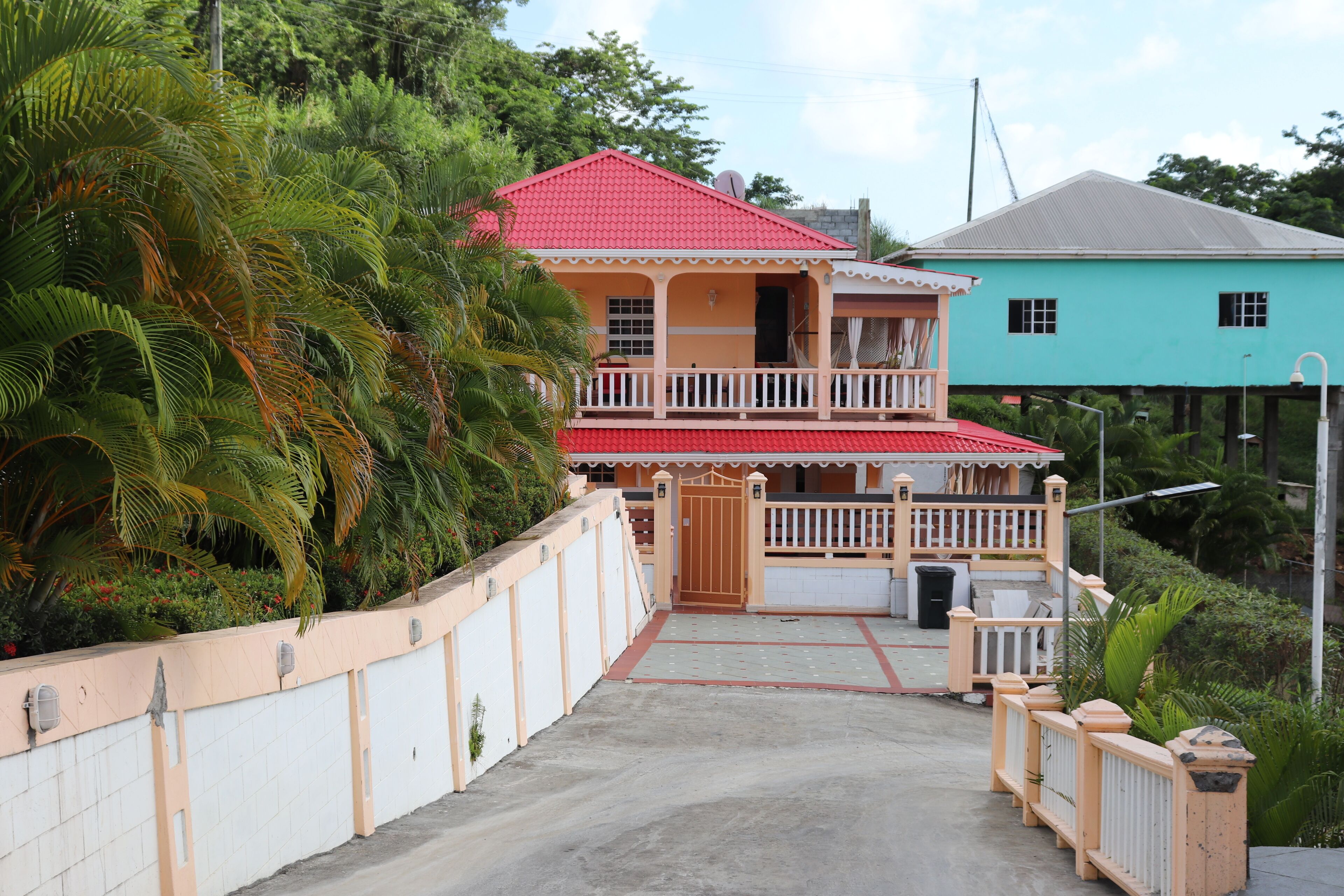 A two-story house with a red roof and a balcony, surrounded by lush greenery.