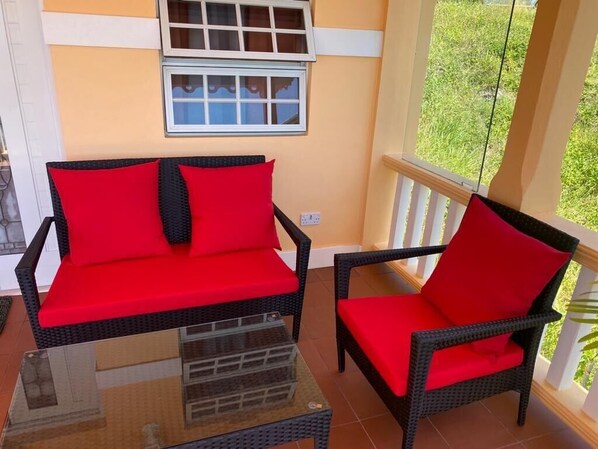 A balcony with wicker furniture and red cushions, a glass table, and a window with white shutters. - Dalys Luxury Apartments (Choppins)
