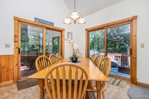 Un comedor con una mesa y sillas de madera, una araña y ventanas amplias con vistas al exterior.