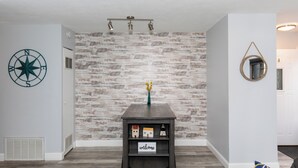 A modern living room with a rustic brick wall, a dark wooden side table with a 'welcome' sign, and a decorative compass on the wall. - Dual Townhomes in Central Downtown Huron (Huron)