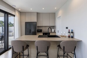 A modern kitchen with a central island, bar stools, a refrigerator, microwave, and wine bottles.