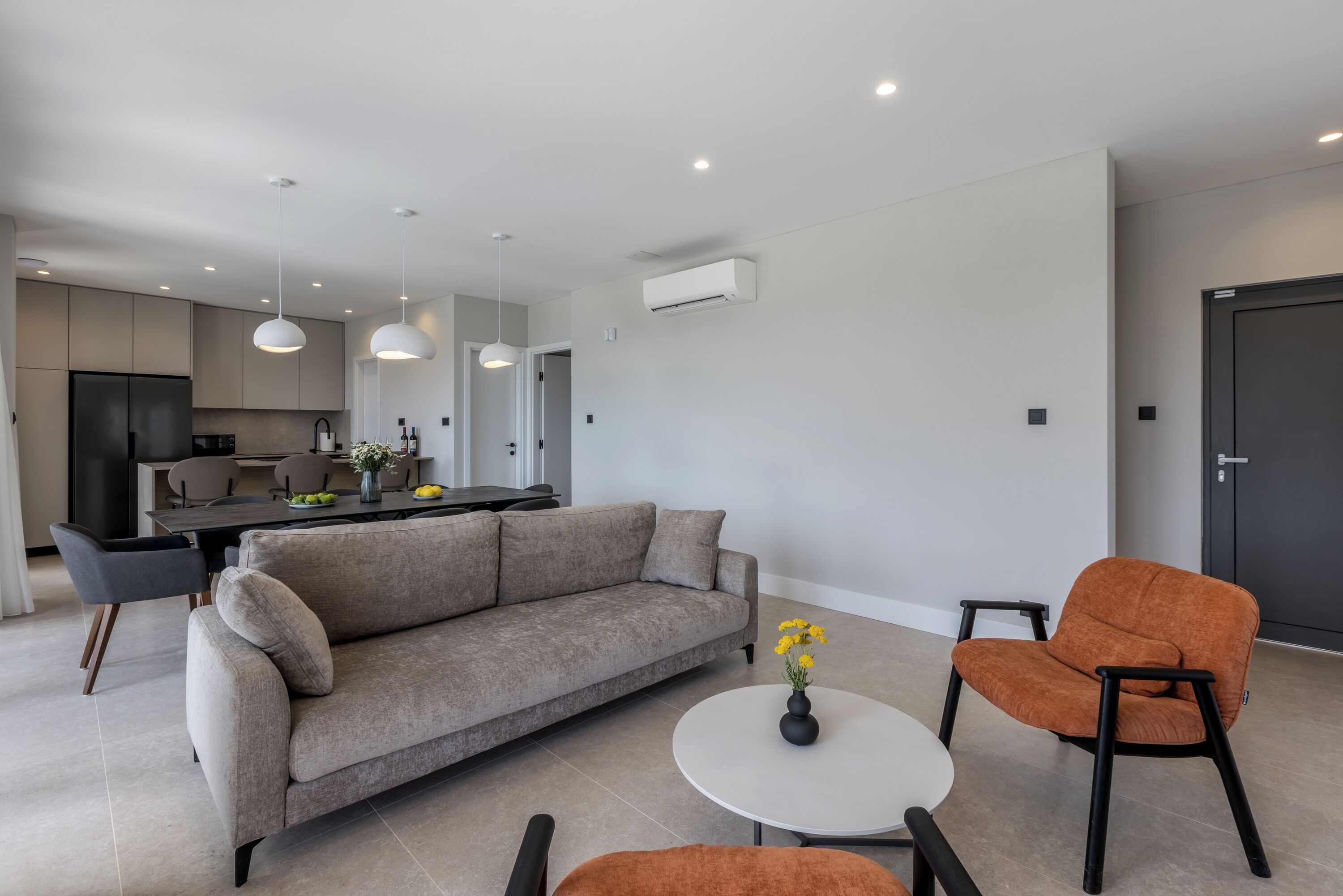 A modern living room with a sofa, a coffee table, and a chair. There is a kitchen in the background with a dining table and chairs.