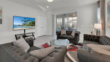 A modern living room with a large flat-screen TV, grey sofas, a glass coffee table, and a chandelier.