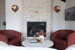 A room with a stone fireplace, two red armchairs, a round coffee table with books and decorative items, and two wall-mounted light fixtures.