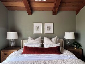 A bedroom with a bed, two nightstands, two lamps, two framed pictures, and a wooden ceiling.