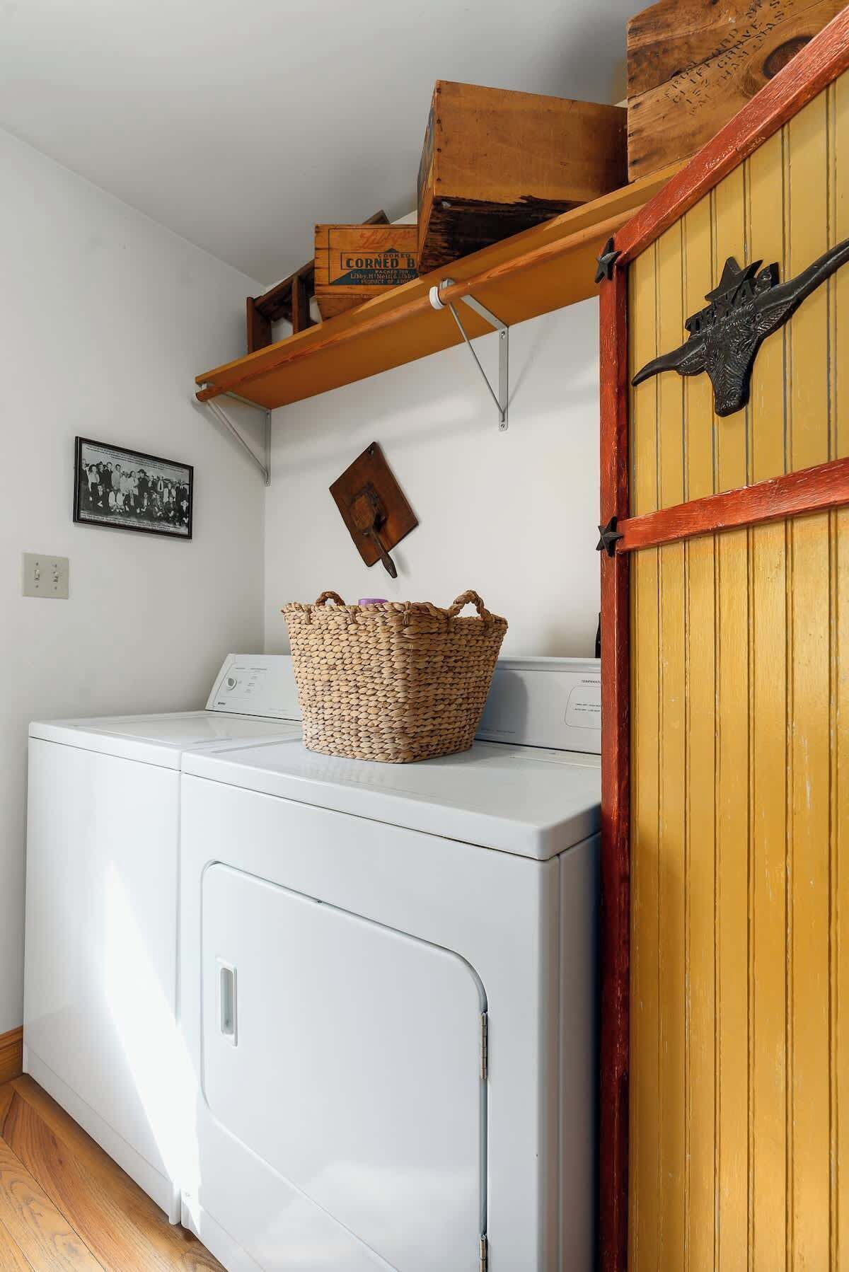 Un espace buanderie avec un lave-linge blanc, un panier en osier posé dessus, une étagère en bois au-dessus, et une porte jaune ornée d’un motif en métal.