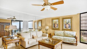 Ein geräumiges Wohnzimmer mit einer großen Couch, einer kleineren Couch, einem Couchtisch und zwei Beistelltischen. Es gibt Deckenventilatoren, ein großes Fenster mit Blick und gerahmte Kunstwerke an den Wänden.