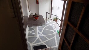 Un couloir étroit, dont le sol est recouvert de carreaux, mène à un balcon. On aperçoit une table avec des plantes en pot sur la gauche.