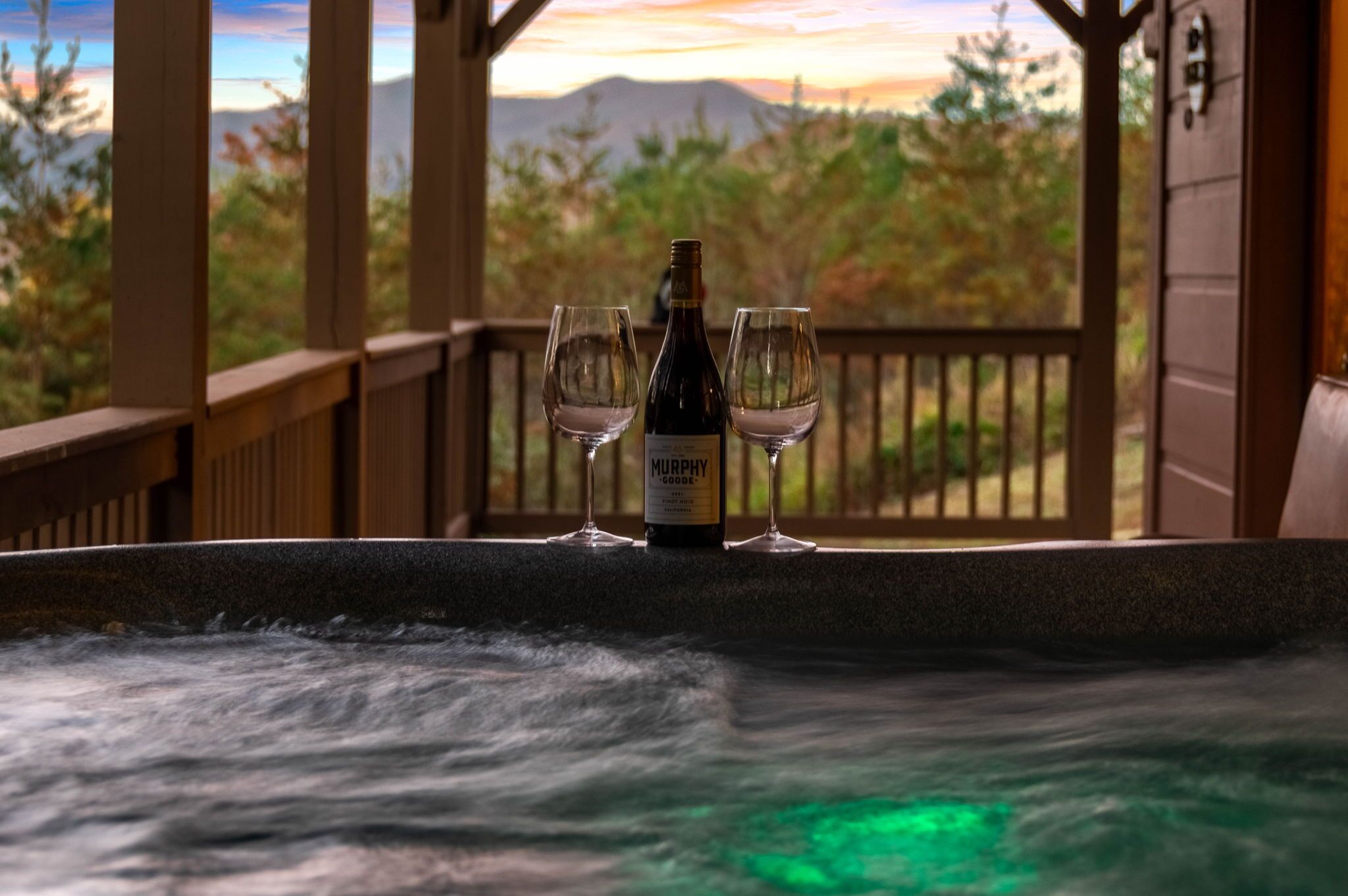 Ein Whirlpool mit einer Flasche Murphy's Grove Wein und zwei Gläsern auf einer Holzterrasse mit Blick auf Berge und Bäume.