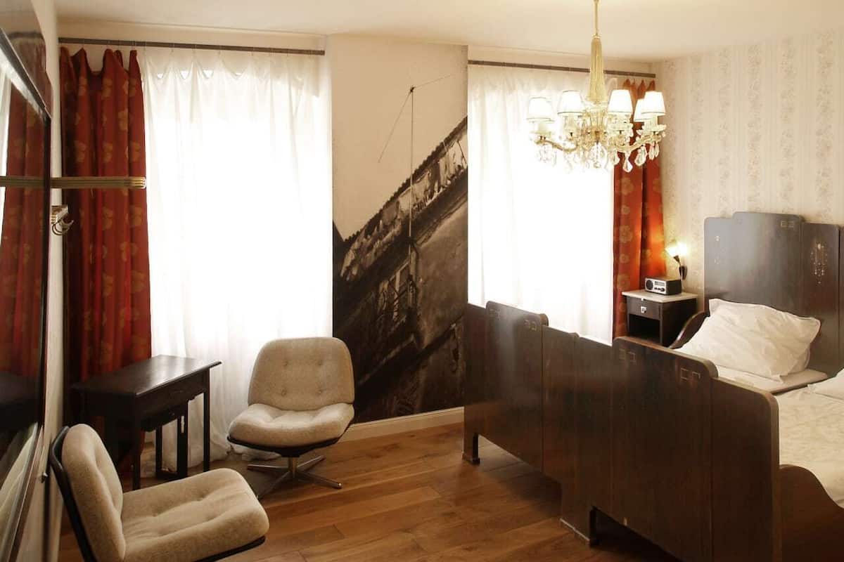 A hotel room with a bed, a chair, a desk, a nightstand, and a chandelier.