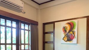 A room with a wooden bench, a small wooden table, a colorful abstract painting on the wall, and a window with curtains. - Hotel Bukit Indah Saumlaki by LeGreen (Saumlaki)