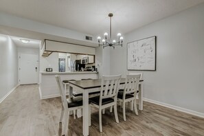 A dining area with a wooden table and chairs, a chandelier, a kitchen in the background, and a framed abstract painting on the wall.