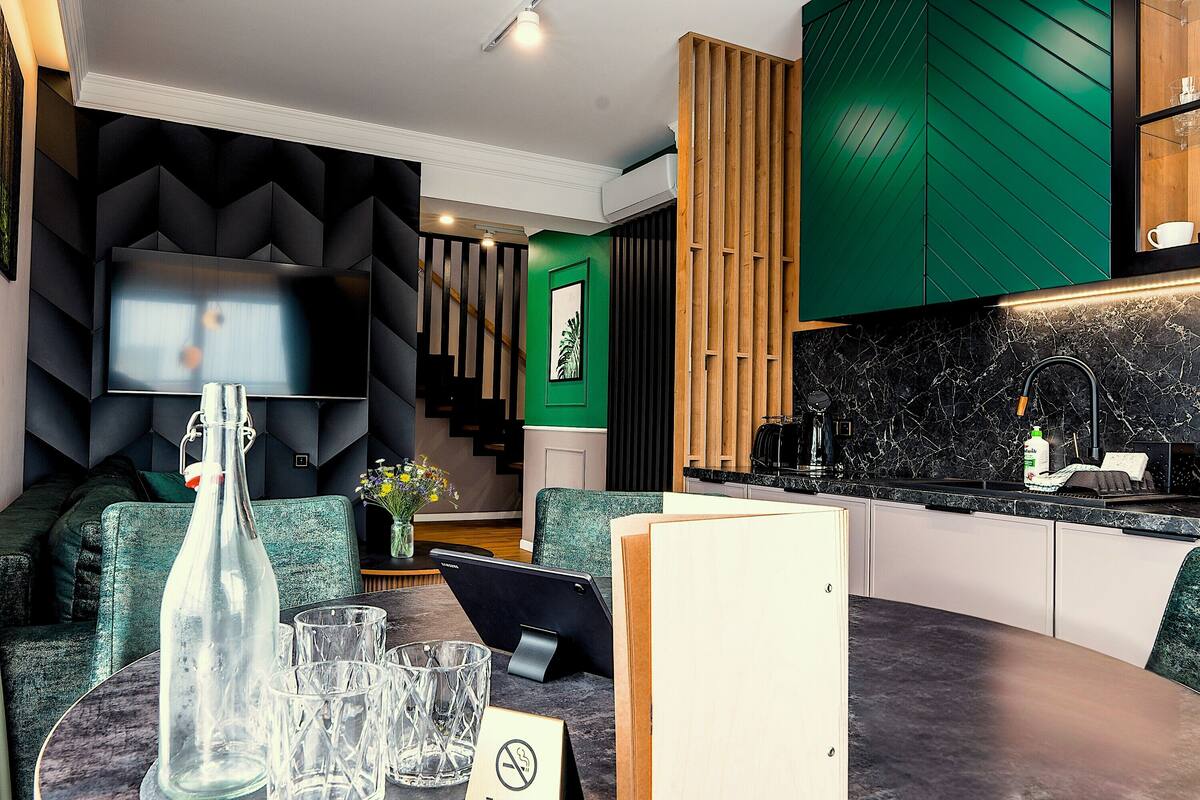 Una sala moderna con un sofá, una mesa de comedor con vasos y una botella, un televisor y una escalera que lleva a una habitación verde.