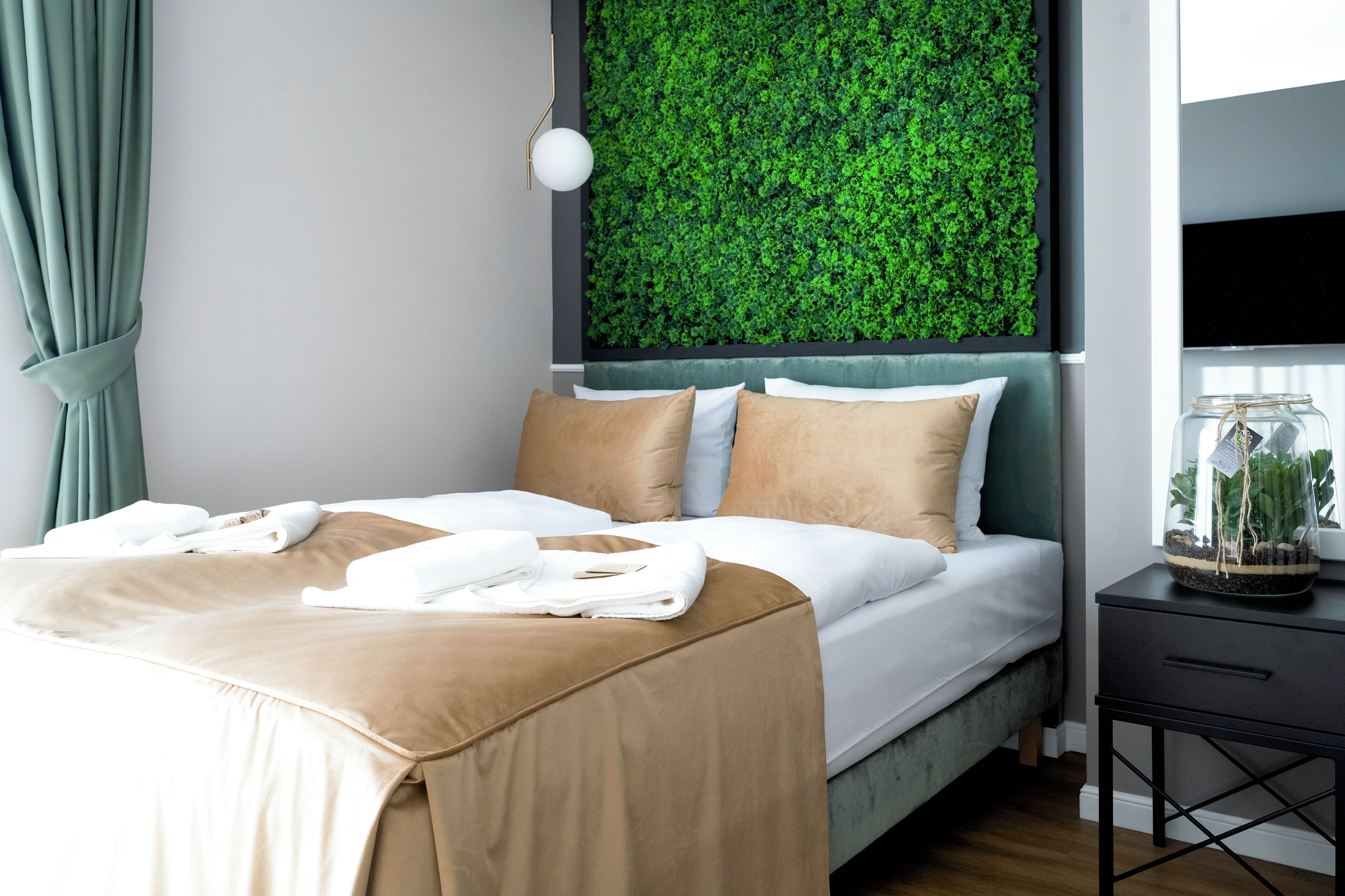 A neatly made bed with a green wall behind it, a small table with a plant, and a window with curtains.