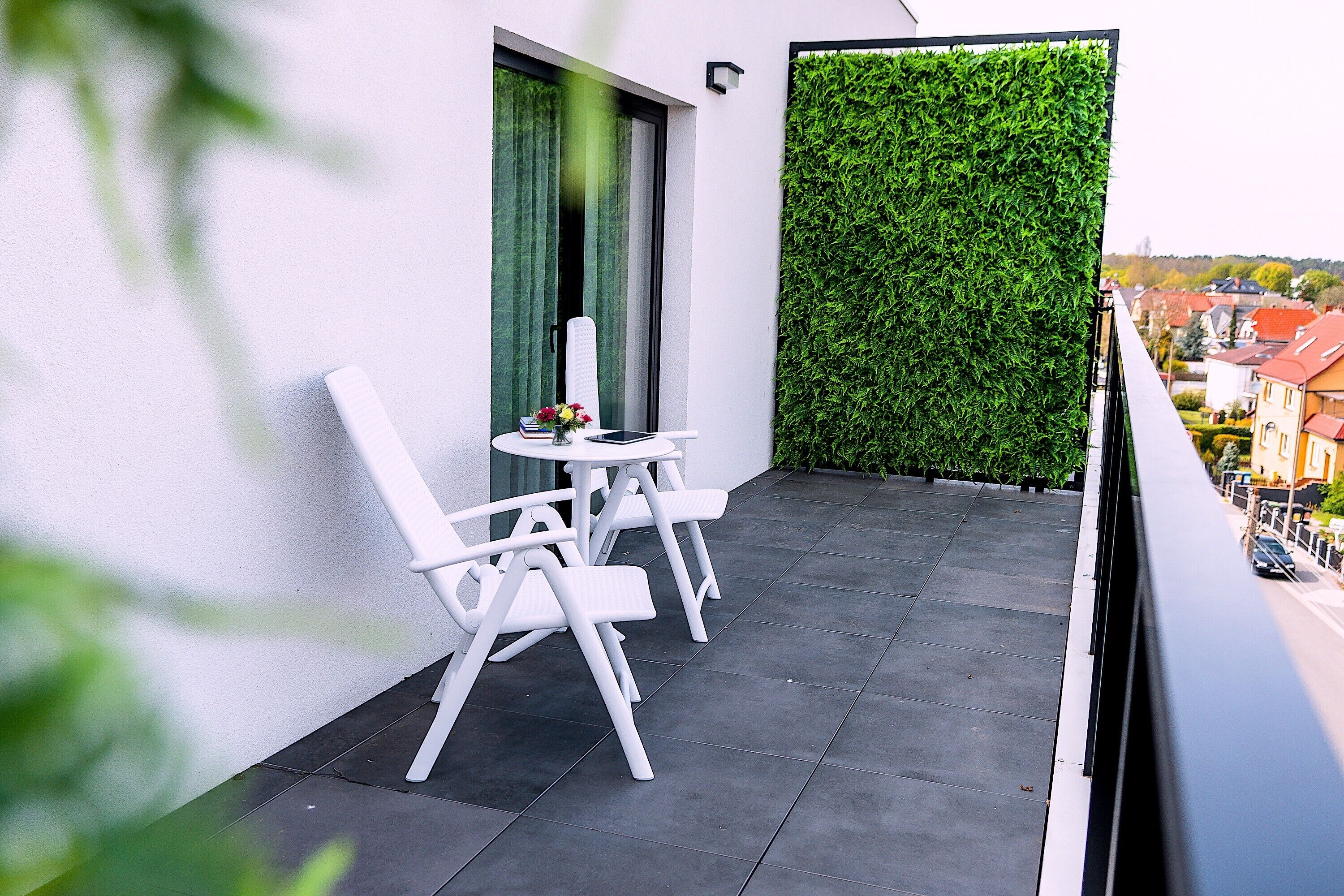 Ein Balkon mit weißen Stühlen und einem Tisch, einer grünen Wand und Blick auf Gebäude.