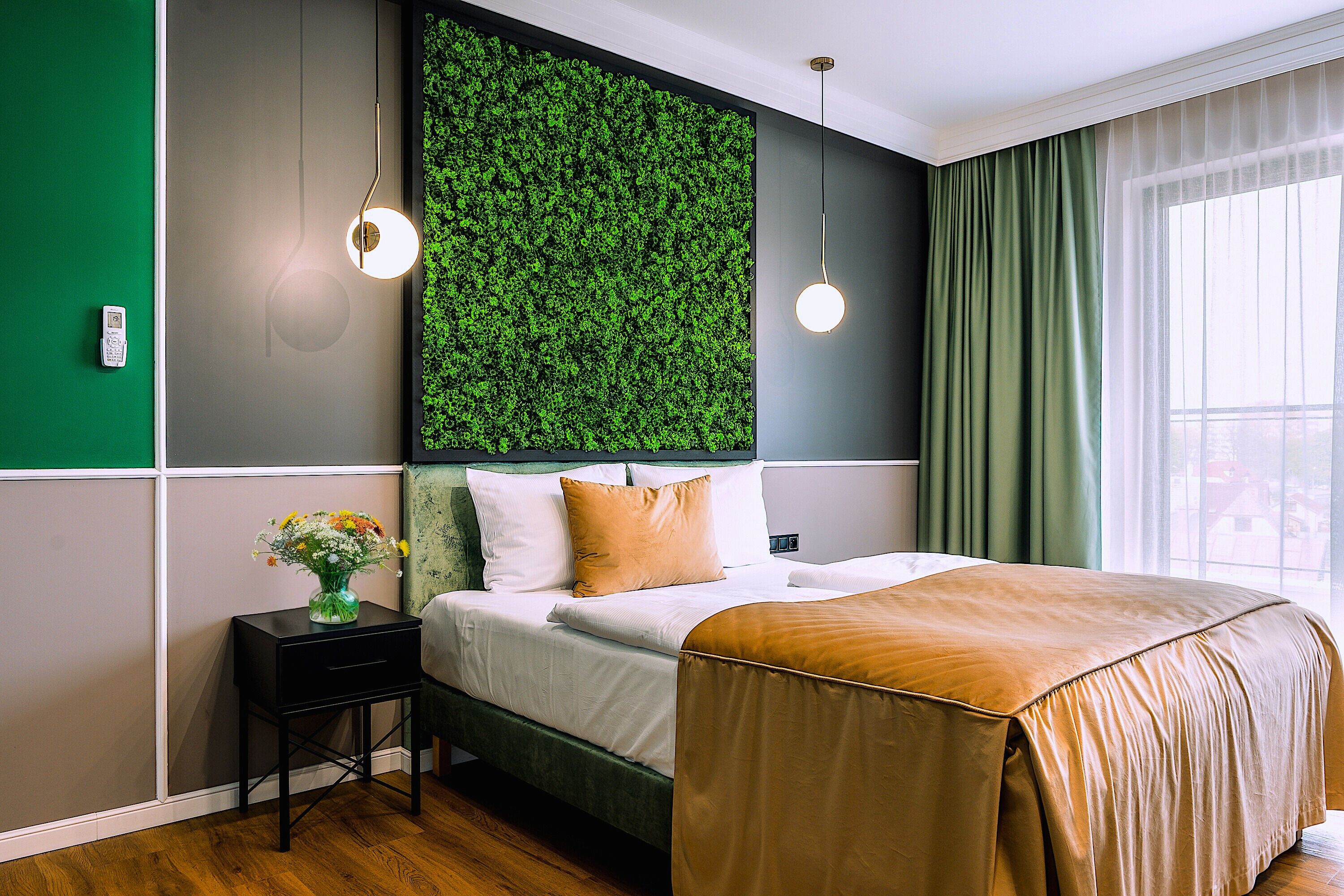 Une chambre d’hôtel moderne avec un mur vert, un lit recouvert d’une courtepointe beige, une table de chevet ornée d’un vase de fleurs et une fenêtre avec des rideaux verts.