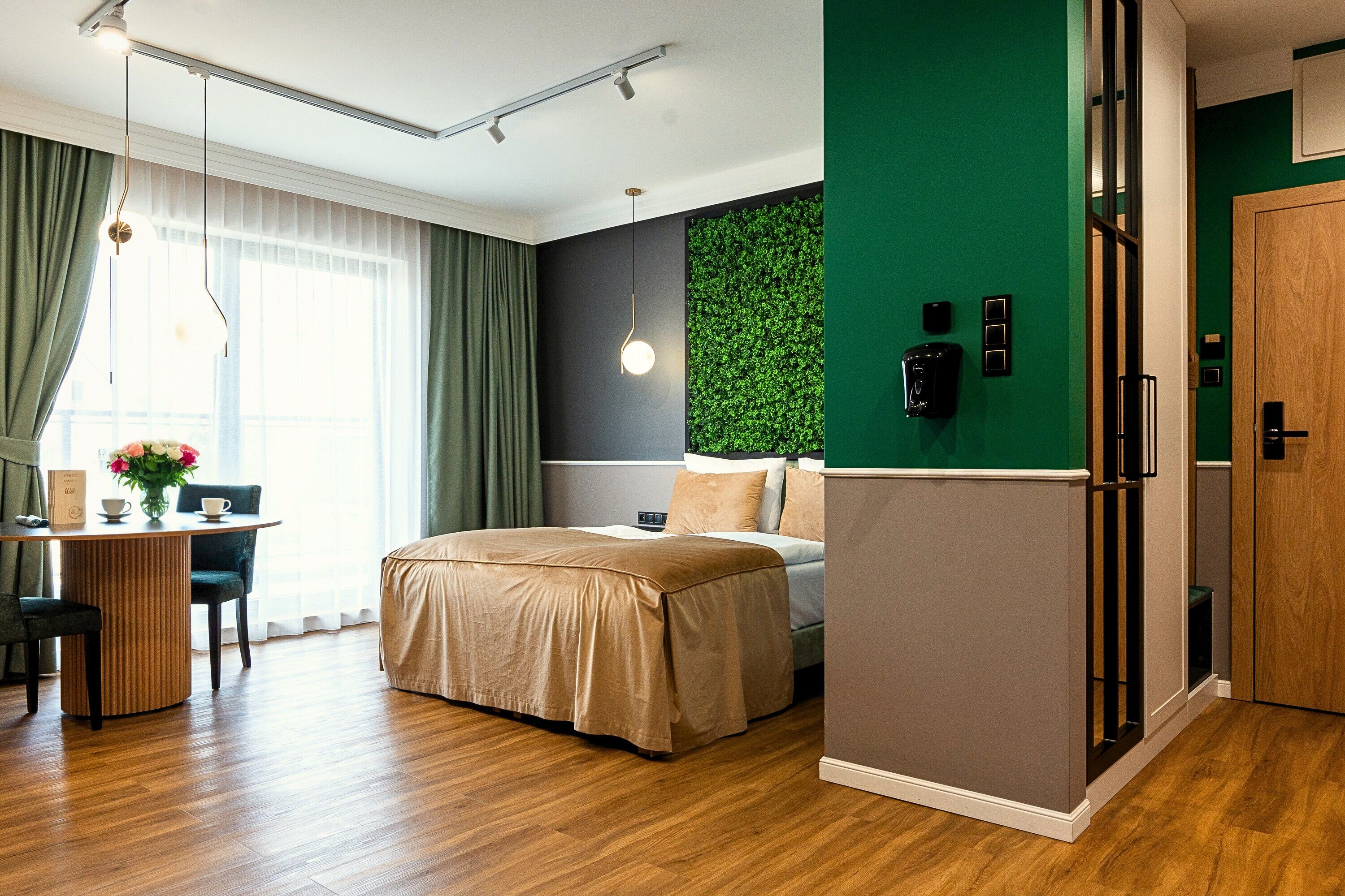 A modern hotel room with a bed, a desk with a chair, a green accent wall, and a wooden door.