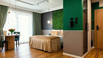 A modern hotel room with a bed, a desk with a chair, a green accent wall, and a wooden door.