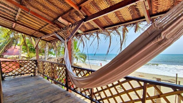 A wooden deck with a hammock, a thatched roof, and a view of the beach and ocean. - Hostal HALALIKU (Santa Marta)