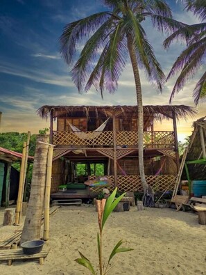 A rustic, two-story wooden structure with a thatched roof, surrounded by palm trees and a sandy ground. - Hostal HALALIKU (Santa Marta)