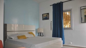 Ett sovrum med en säng, ett sängbord, en lampa, ett fönster med gardiner och en tavla som hänger på väggen.