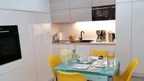Una cocina moderna con armarios blancos, un horno negro, un lavamanos de acero inoxidable y una cafetera. Hay una mesa de vidrio con sillas amarillas y un florero.