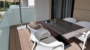 Un área de comedor al aire libre con un diseño moderno, que incluye una mesa de madera, sillas de mimbre blanco, una división de vidrio y un paraguas grande.