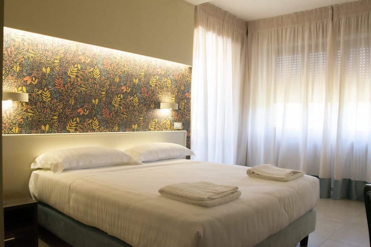 A hotel room with a large bed, two bedside lamps, a floral-patterned headboard, and sheer curtains.