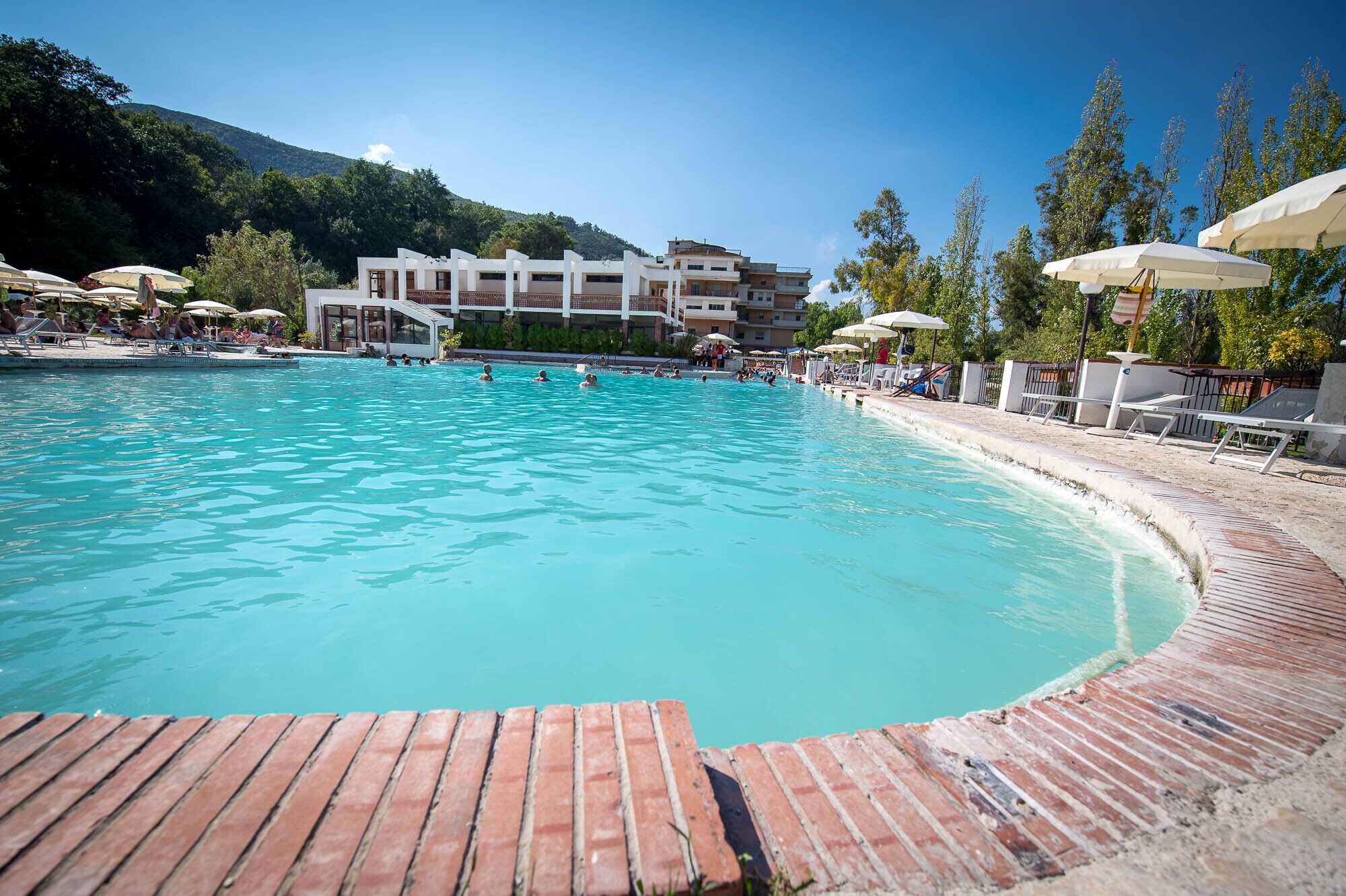 Ein Schwimmbecken mit Holzterrasse im Vordergrund, umgeben von Liegestühlen und Sonnenschirmen.