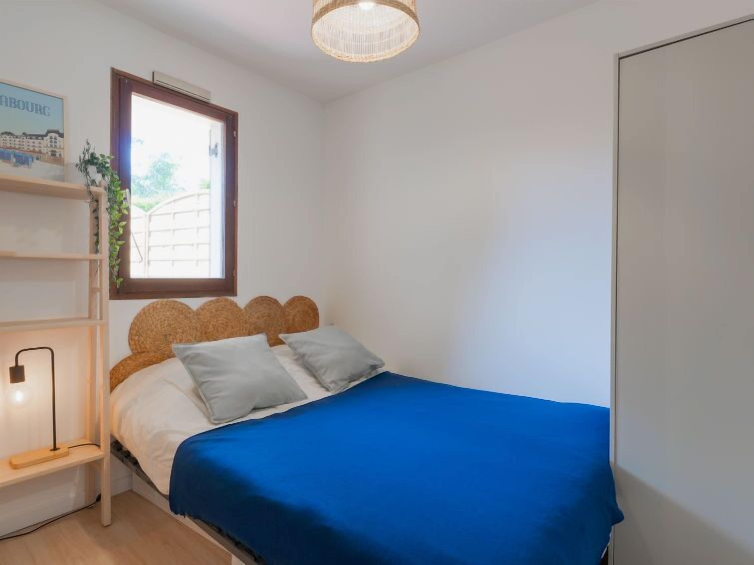 Quarto com cama, mesa de cabeceira, luminária, janela com vista e prateleira na parede com uma planta.