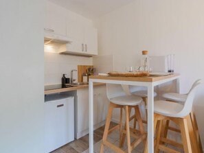 Eine kleine Küche mit weißen Schränken, einem Kühlschrank, einem Holztisch und zwei weißen Barhocker.