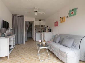 Un espace de vie compact comprenant un coin cuisine, un canapé gris, une petite table avec une bouteille et un verre, et un ventilateur de plafond.