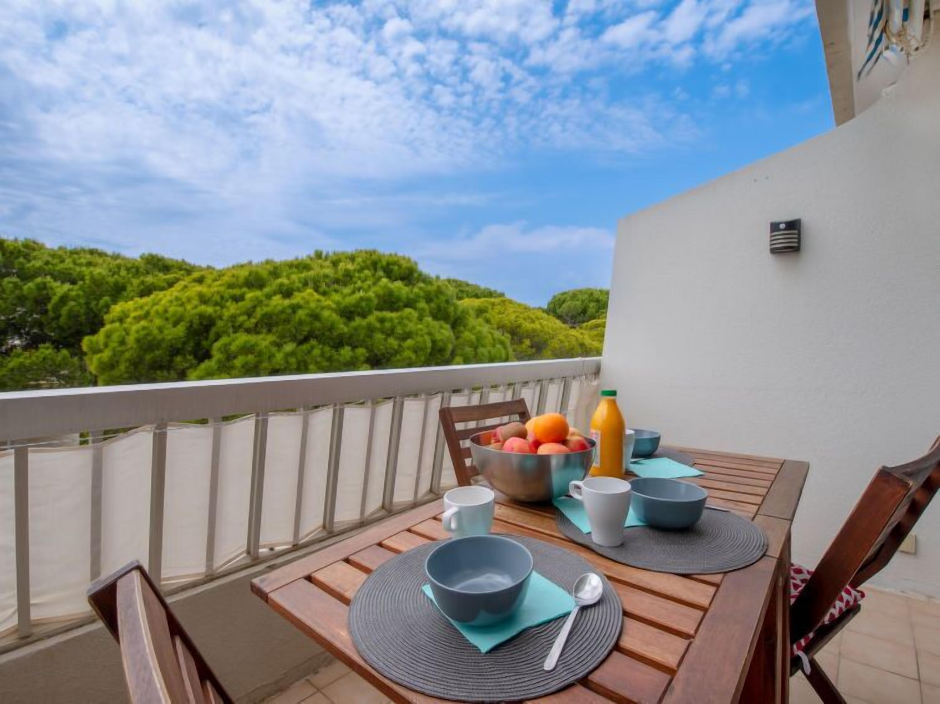 A wooden table set with bowls, cups, and a fruit bowl on a balcony with a view of green trees and a clear sky.