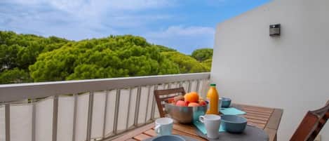 A wooden table set with bowls, cups, and a fruit bowl on a balcony with a view of green trees and a clear sky.