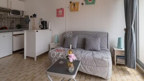 Ett kompakt vardagsrum med ett litet pentry, en grå soffa, ett litet bord med en flaska och blommor samt ett fönster med gardiner.