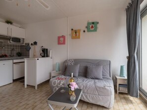 Un espace de vie compact comprenant un coin cuisine, un canapé gris, une petite table avec une bouteille et des fleurs, et une fenêtre avec des rideaux.
