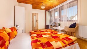 Un dormitorio con techo de madera, una cama grande con colcha colorida, una mesita, una silla y una ventana con cortinas translúcidas.