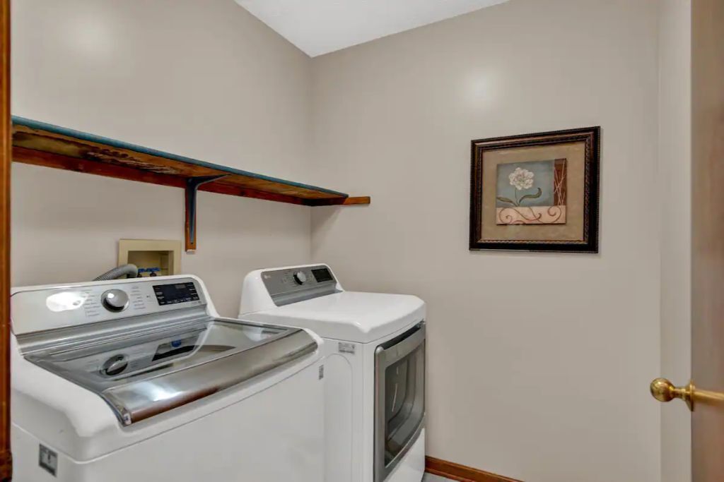 A laundry room with a front-loading washer and dryer, a wooden shelf above the dryer, and a framed picture on the wall.