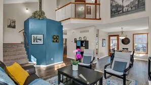 A spacious living room with a blue accent wall, a staircase leading to a balcony, and a dining area with a chandelier. - Entire 3-level, 3 BRM home w/King/Queen/Twin beds (Topeka)