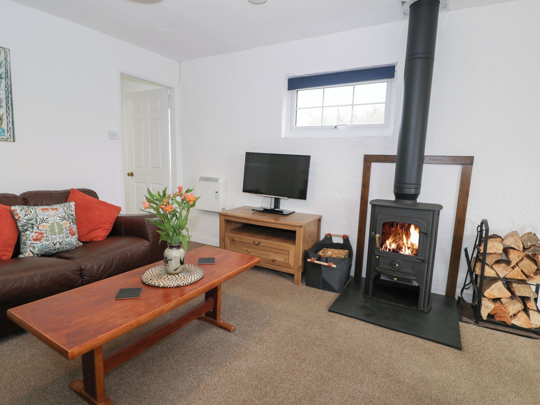 A living room with a brown leather sofa, a wooden coffee table, a TV on a wooden stand, and a black wood-burning stove.