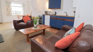 A living room with a brown leather sofa, a wooden coffee table, and a small table with a vase of flowers. There is a kitchen area with blue cabinets and a white refrigerator. - Min Y Mor (Criccieth)