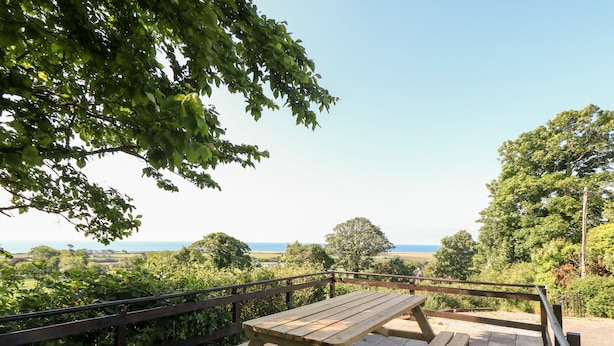 A wooden picnic table on a paved deck with a metal railing, overlooking a scenic landscape with trees and a distant view of the sea. - Min Y Mor (Criccieth)