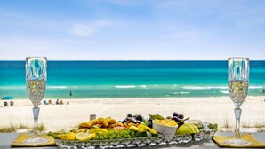 Een tafel met een plateau visgerechten en twee glazen witte wijn op een strand, met een helderblauwe lucht en de oceaan op de achtergrond.