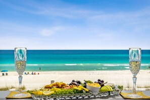 A table with a seafood platter and two glasses of white wine on a beach with a clear blue sky and ocean in the background.