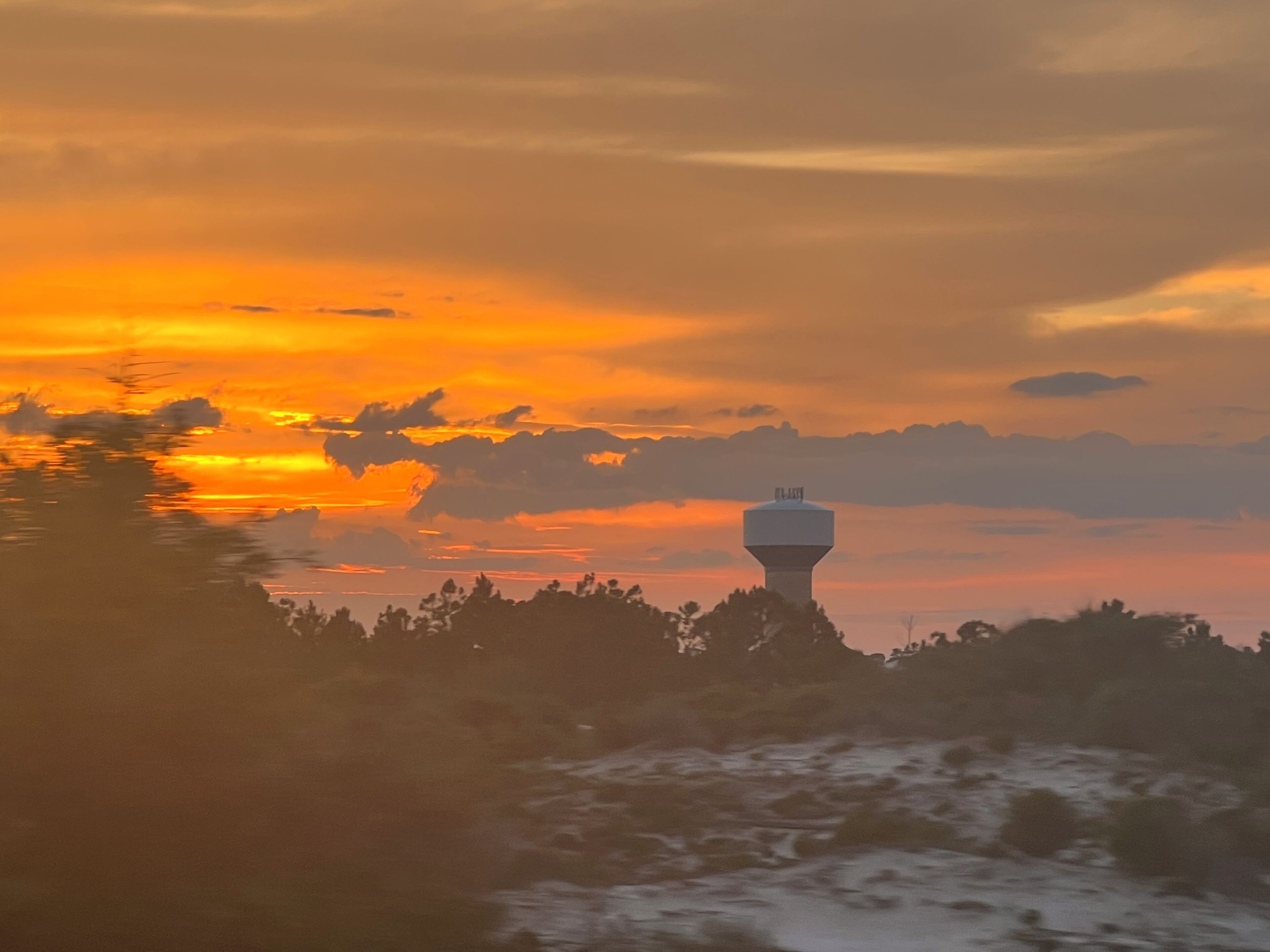 A water tower with a flat top, surrounded by trees and a body of water reflecting the sunset.