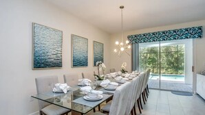 Un comedor con una mesa de vidrio, sillas blancas y vistas a una piscina a través de grandes puertas correderas de cristal.