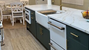 Eine moderne Küche mit einer zentralen Kochinsel, weißen Schränken, dunkelgrünen Unterschränken und goldenen Griffen. Zwei Pendelleuchten mit mehreren Lampen, eine weiße Arbeitsplatte und ein Holzschneidebrett auf der Insel.